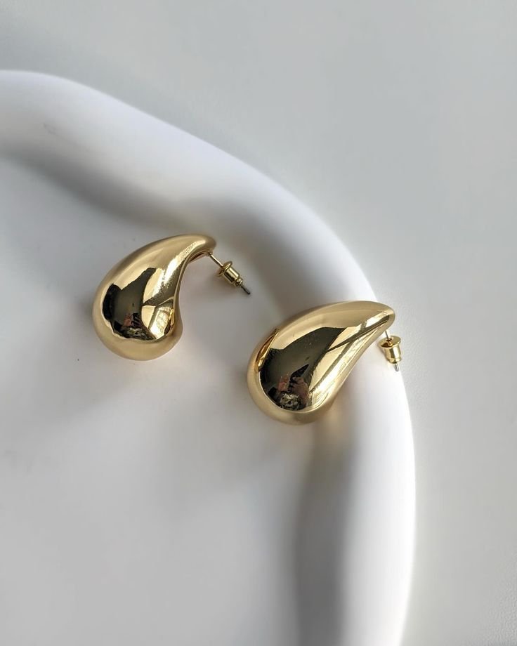Gold teardrop-shaped stud earrings with posts resting on a white plate surface.