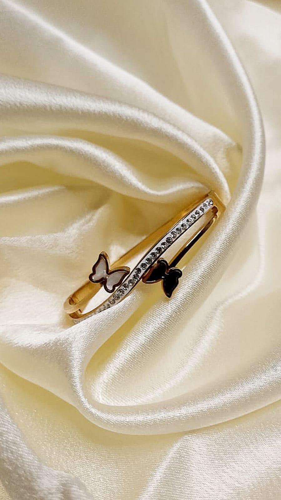 Gold bracelet with a row of small diamonds and two butterfly charms, one white and one black, resting on cream satin.