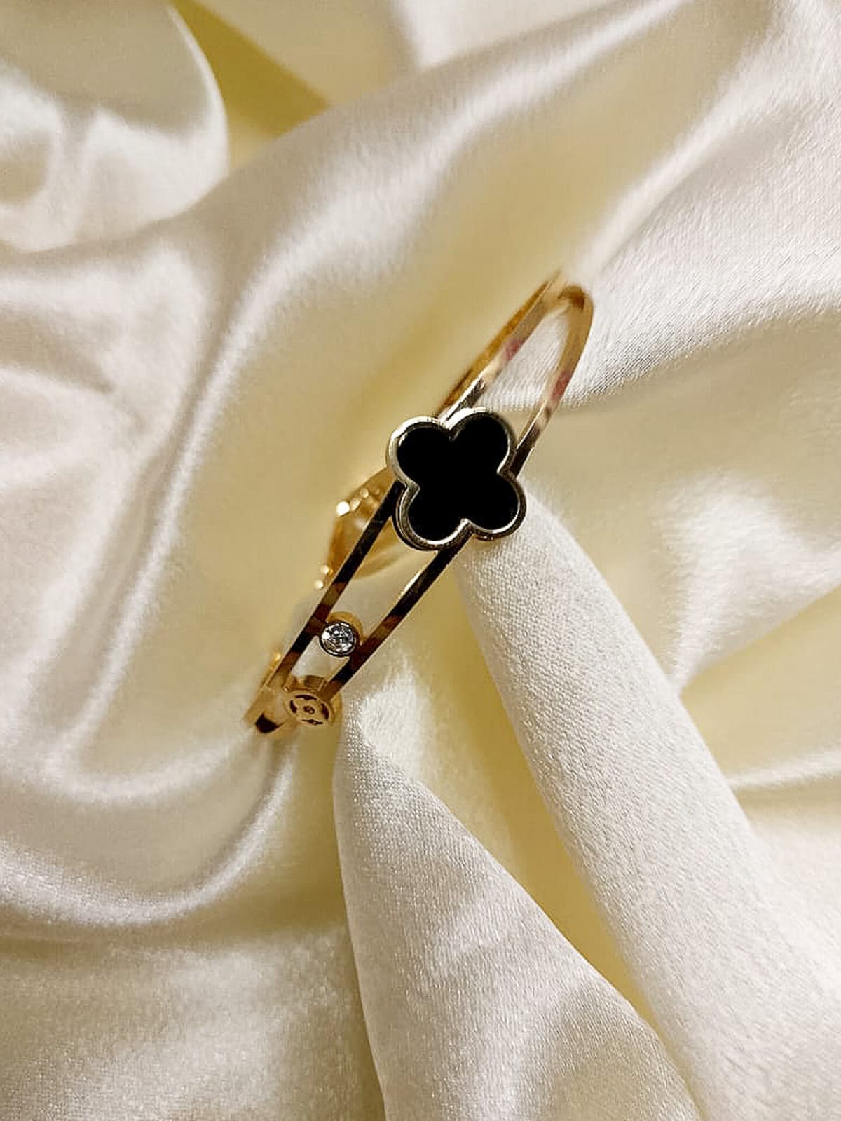 Gold bracelet with a black four-leaf-clover charm resting on ivory satin fabric, with a small diamond accent nearby.