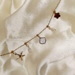 Delicate gold chain necklace with charms: starfish, clover, pearl, and pale gemstone on cream satin fabric, positioned diagonally.