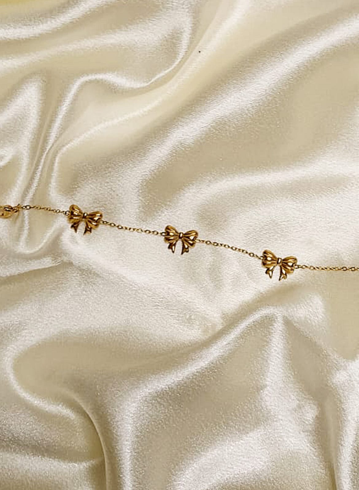 Delicate gold chain bracelet with small bow charms on ivory satin backdrop.