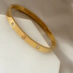 Gold bracelet with small diamond accents resting on a white surface, showing screw-like details along the band.