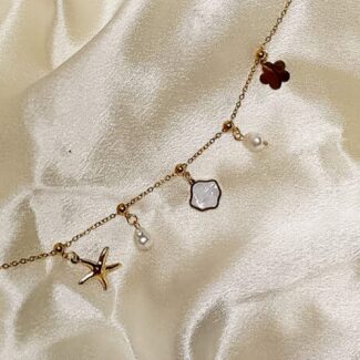 Delicate gold chain necklace with charms: starfish, clover, pearl, and pale gemstone on cream satin fabric, positioned diagonally.