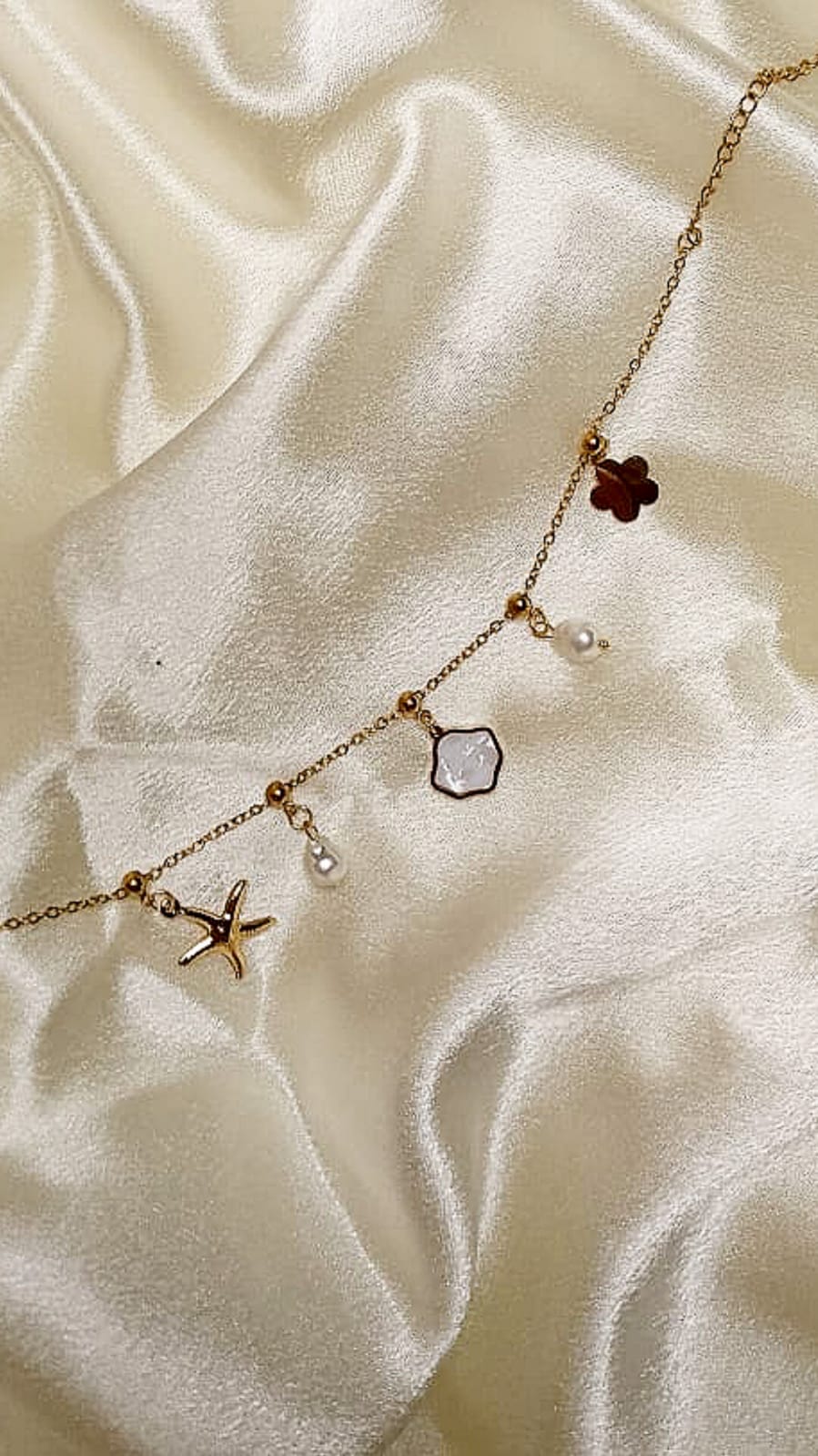 Delicate gold chain necklace with charms: starfish, clover, pearl, and pale gemstone on cream satin fabric, positioned diagonally.