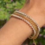 Wrist wearing multiple stacked gold and diamond tennis bracelets with clear stones gleaming in sunlight.