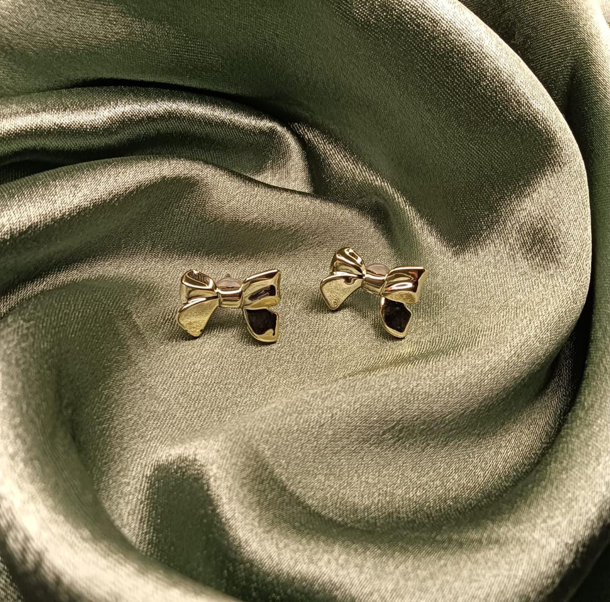 Gold bow-shaped stud earrings resting on emerald green satin fabric