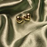 Two small gold heart earrings resting on green satin fabric, gleaming in the light.