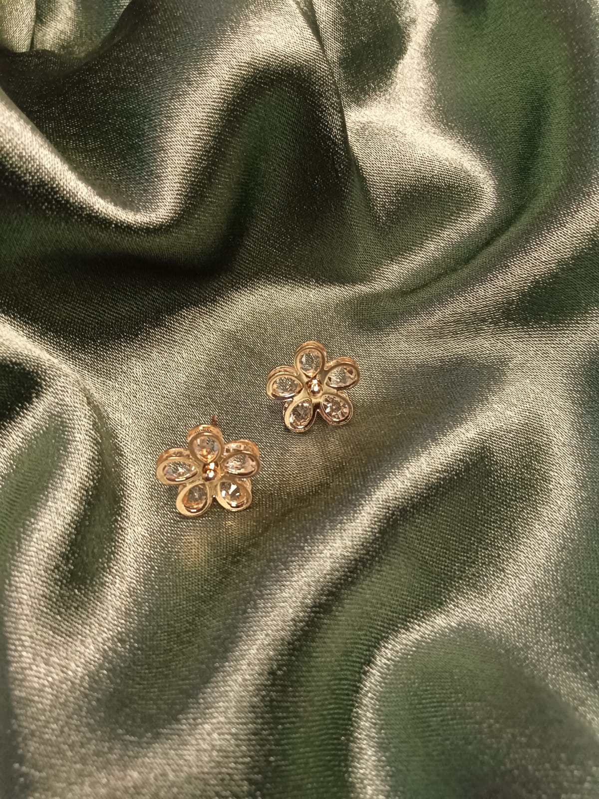 Gold-tone flower-shaped earrings with clear gemstones, placed on dark green satin fabric.