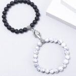 Two beaded bracelets laid on a white surface: one with black matte beads and a silver praying-hands charm, the other with white marble-pattern beads.