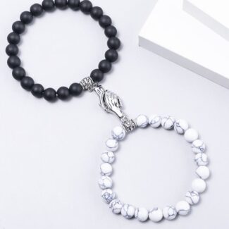 Two beaded bracelets laid on a white surface: one with black matte beads and a silver praying-hands charm, the other with white marble-pattern beads.