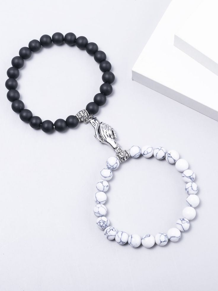 Two beaded bracelets laid on a white surface: one with black matte beads and a silver praying-hands charm, the other with white marble-pattern beads.