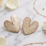 Two heart-shaped earrings with iridescent cream interiors and rose-gold edges, resting on a marble surface.