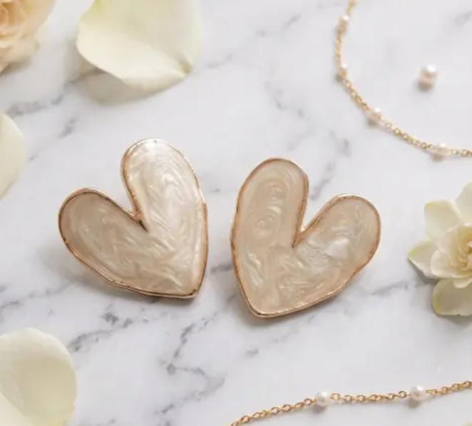 Two heart-shaped earrings with iridescent cream interiors and rose-gold edges, resting on a marble surface.