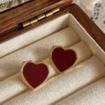 Gold heart-shaped enamel earrings in a wooden jewelry box, resting on a light fabric lining.