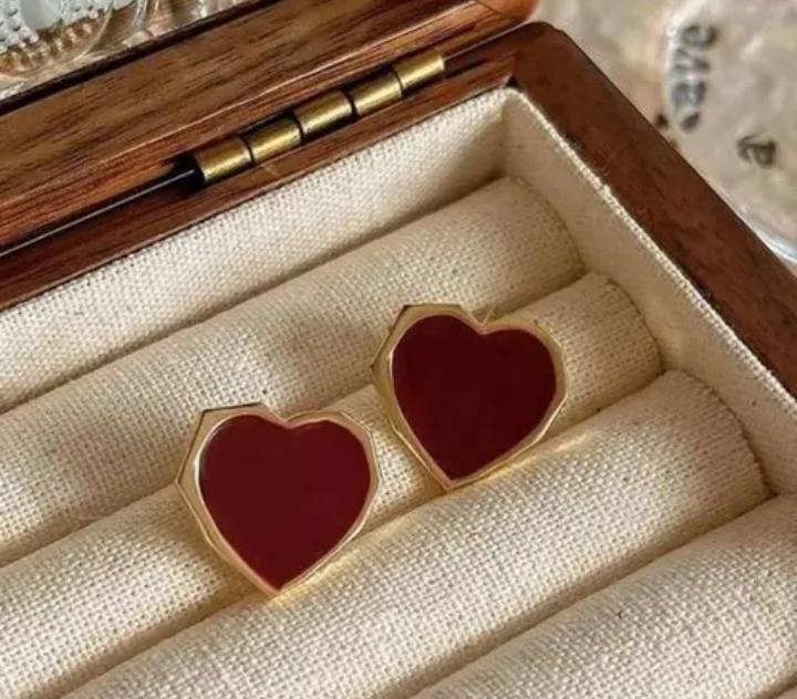 Gold heart-shaped enamel earrings in a wooden jewelry box, resting on a light fabric lining.