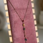 Copper-toned necklace with a row of small colored gems, displayed on a pink velvet stand with a wooden frame.