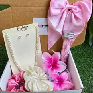 Gift box containing pink fashion items: pearl necklace, earrings, satin bow, plumeria flowers, and a small pink bottle in a carded display.