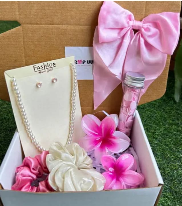 Gift box containing pink fashion items: pearl necklace, earrings, satin bow, plumeria flowers, and a small pink bottle in a carded display.