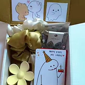Open gift box with cartoon drawings on the back wall, a yellow paper flower, and a birthday card showing a party-hatted ghost holding a cake.