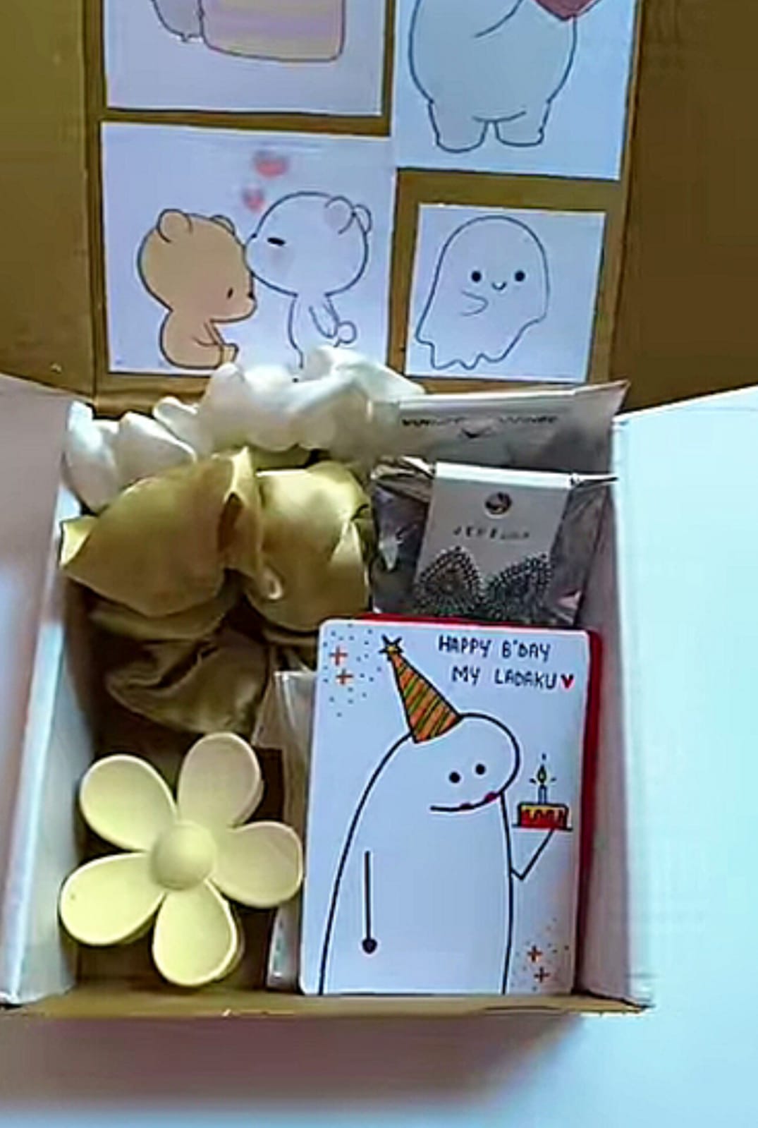 WhatsApp Image 2026-04-25 at 12.58.02 (1) Open gift box with cartoon drawings on the back wall, a yellow paper flower, and a birthday card showing a party-hatted ghost holding a cake.