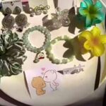 Assorted jewelry pieces on a white display tray, including earrings, bracelets, and oversized flower clips with a cute cartoon card in front.