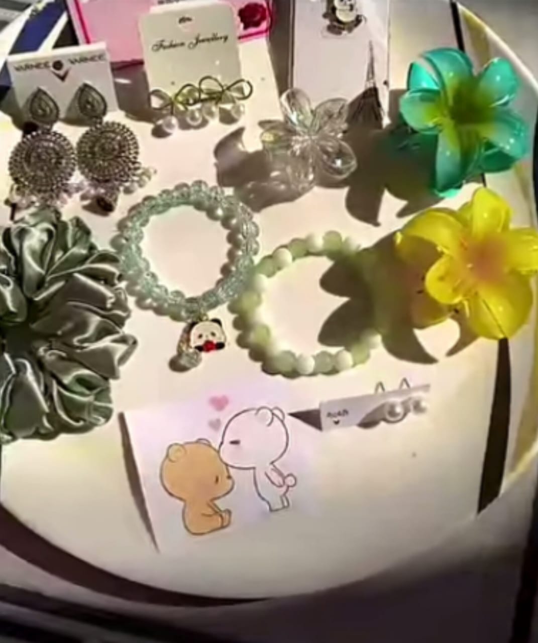 Assorted jewelry pieces on a white display tray, including earrings, bracelets, and oversized flower clips with a cute cartoon card in front.