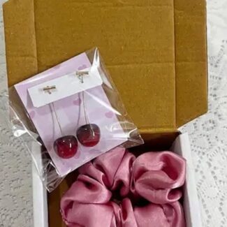 Open cardboard box with pink fabric scrunchie and two flower-shaped soaps; small cherry earrings in plastic packaging on top.