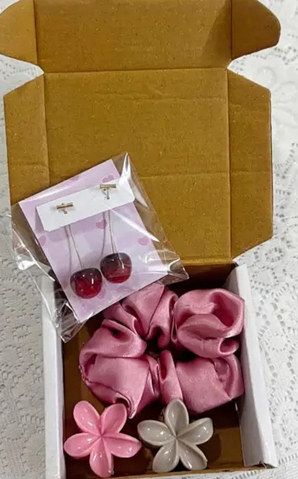 Open cardboard box with pink fabric scrunchie and two flower-shaped soaps; small cherry earrings in plastic packaging on top.