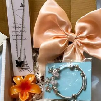 Gift box interior showing a peach satin bow, orange flower, and jewelry cards including a butterfly necklace with an Eiffel Tower design card on the left and a blue bracelet card on the right.