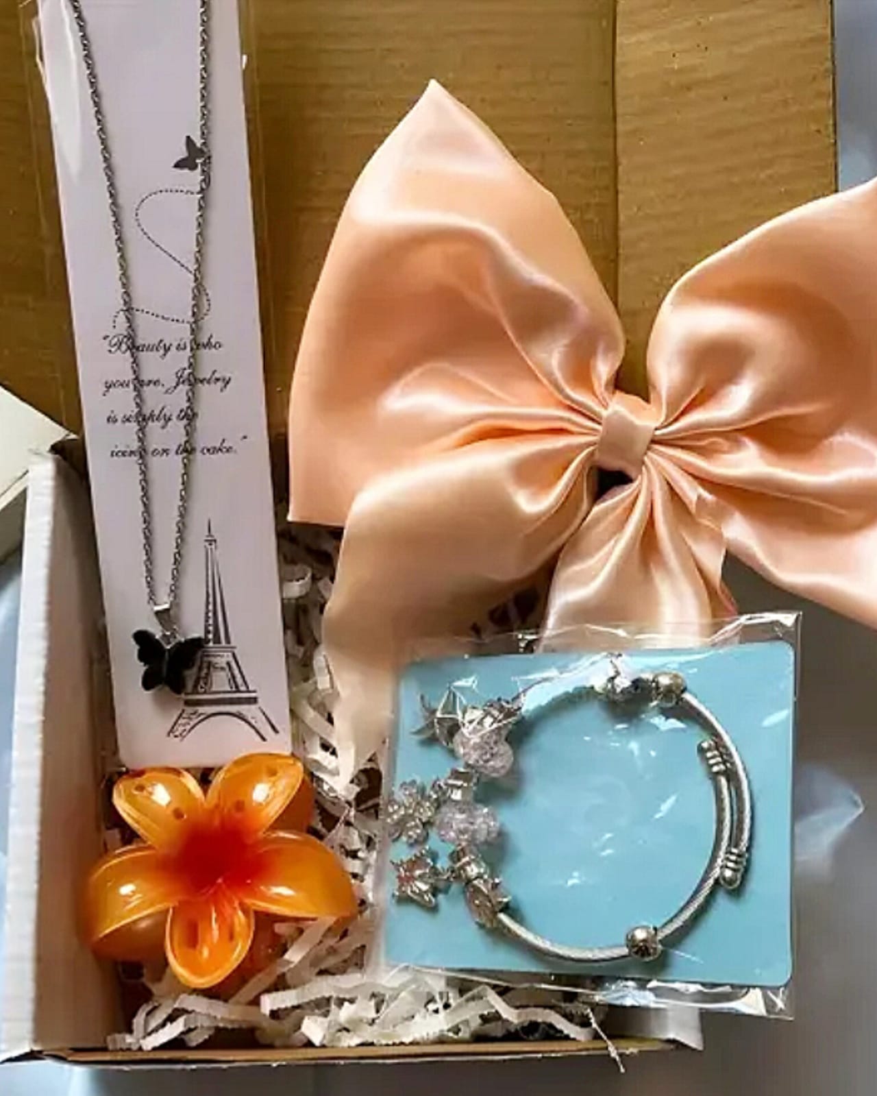 Gift box interior showing a peach satin bow, orange flower, and jewelry cards including a butterfly necklace with an Eiffel Tower design card on the left and a blue bracelet card on the right.