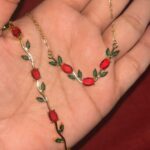 Gold-tone necklace shaped like a vine with red oval gems and green leaf accents held in a hand.