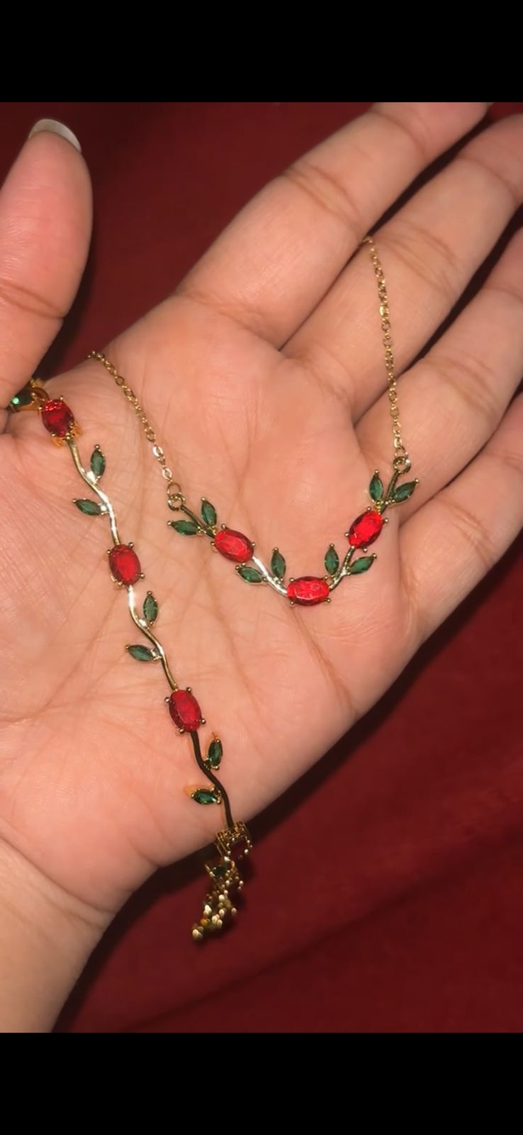 Gold-tone necklace shaped like a vine with red oval gems and green leaf accents held in a hand.