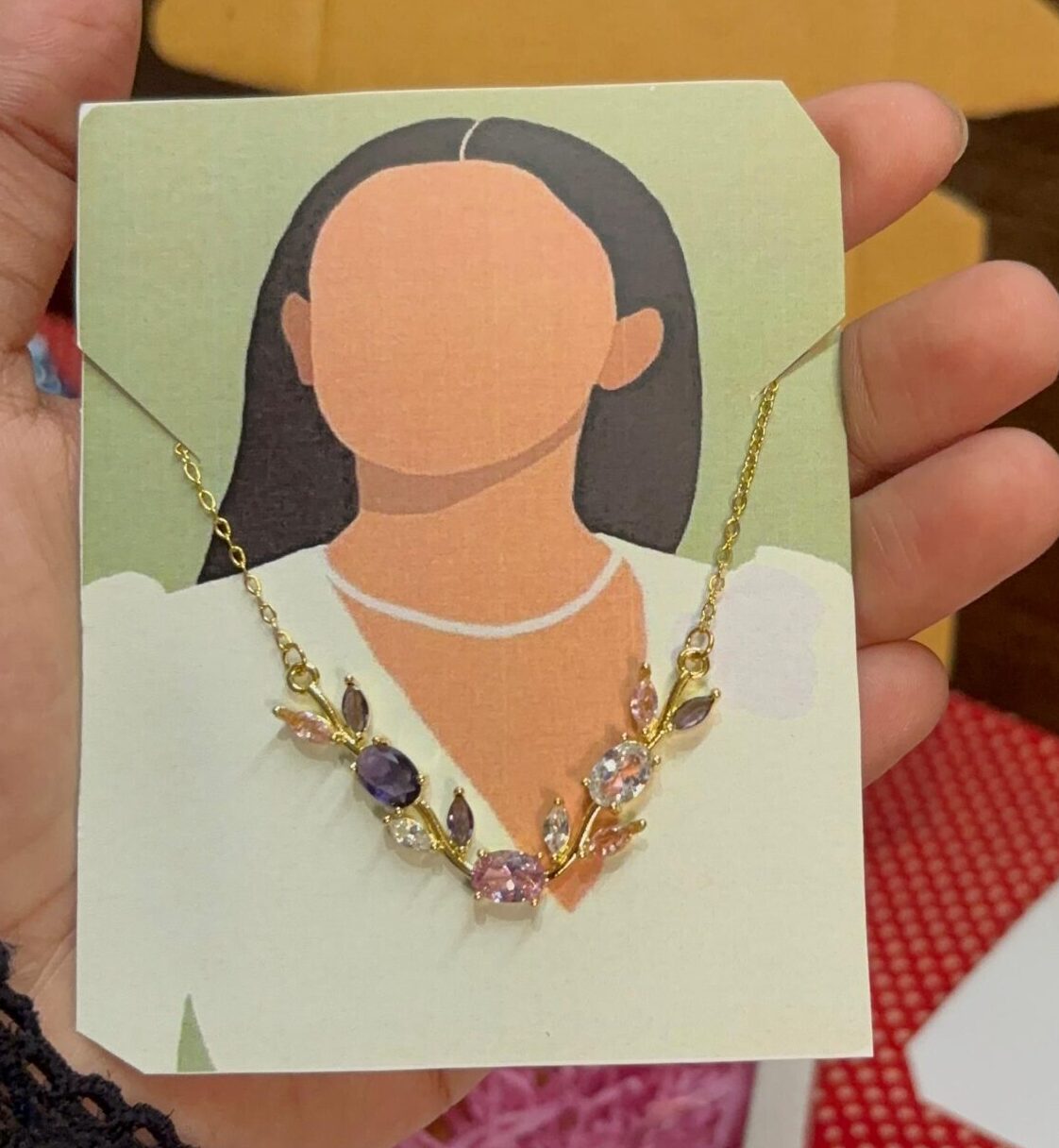 Illustrated jewelry card: a faceless figure wearing a gold necklace with pink, purple, and clear stones.