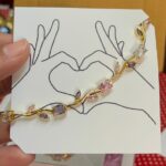 Gold-tone bracelet with pink and clear stones resting on a white card featuring a hand-drawn heart formed by two hands.