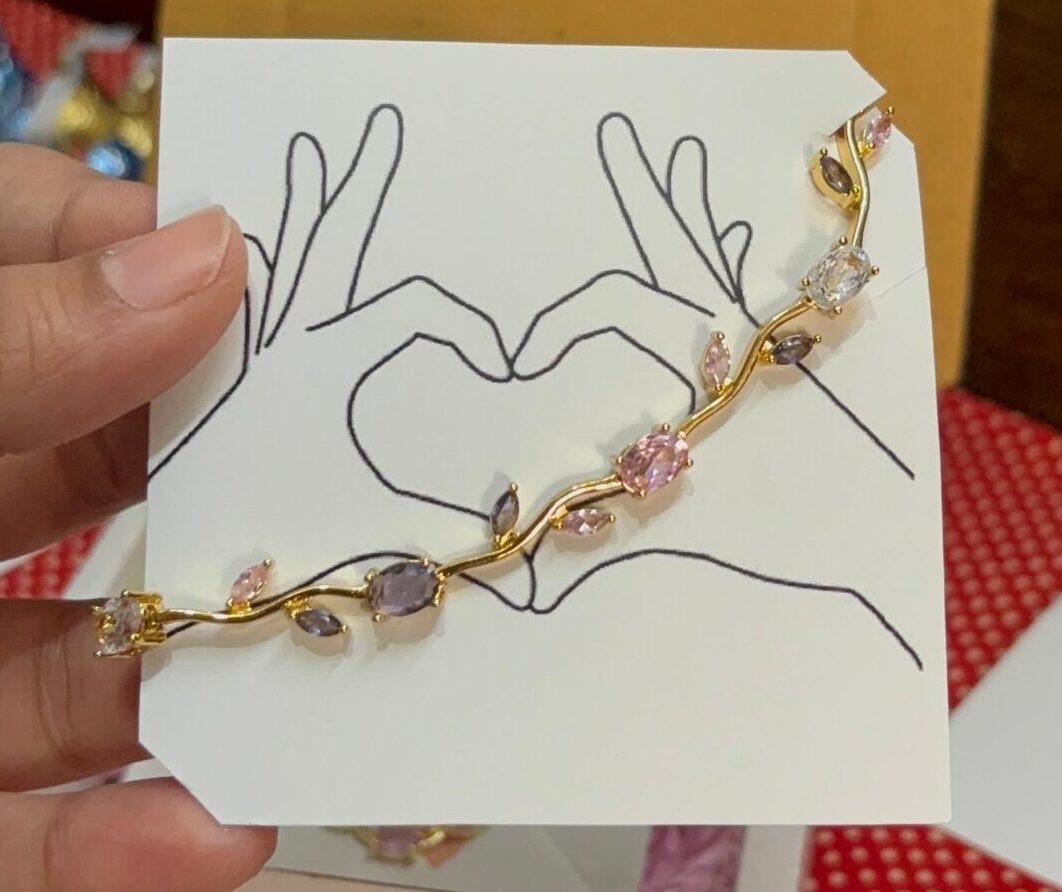 Gold-tone bracelet with pink and clear stones resting on a white card featuring a hand-drawn heart formed by two hands.