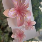 Pink plumeria flower hair clip on white backing held by a hand outdoors.
