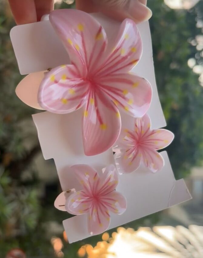 Pink plumeria flower hair clip on white backing held by a hand outdoors.