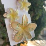 Three pale yellow plumeria flowers with turquoise speckles mounted on a white display card, held by a hand outdoors.