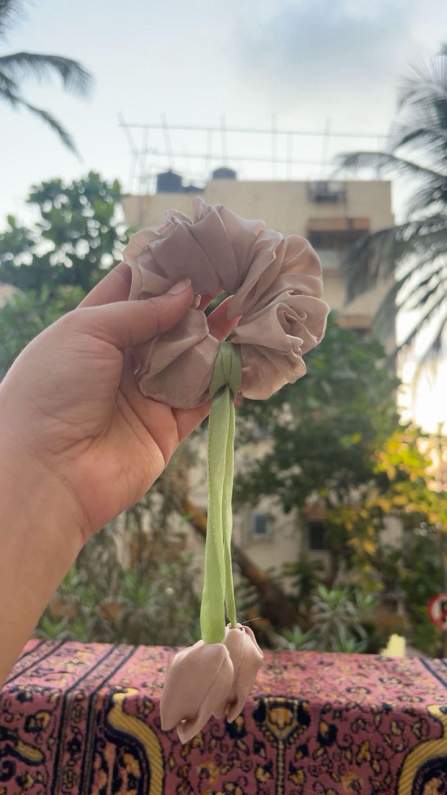 Beige fabric flower scrunchie on a green ribbon held by a hand, outdoors with trees and a building in the background.