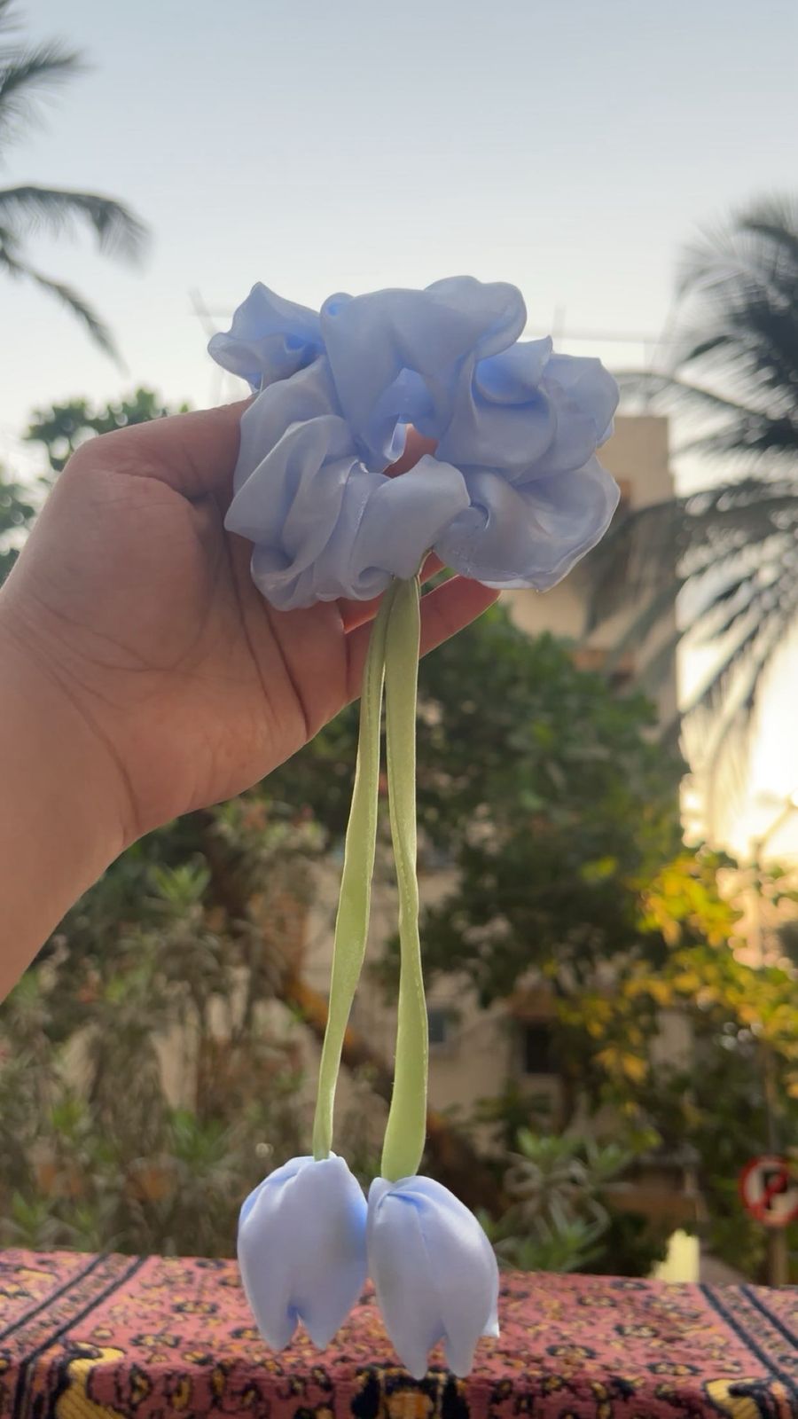 Hand holding a light-blue fabric flower hair scrunchie with two long pale green ribbons, outdoors with palm trees in the background.