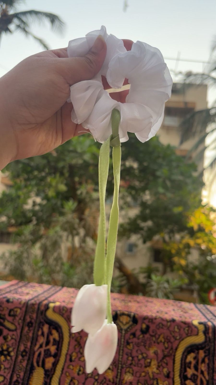 Hand holding a white fabric scrunchie tied around a long green stem with two hanging white petals, outdoors on a patterned surface in the background.