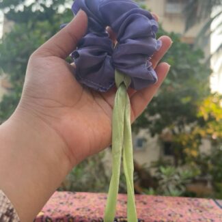 Purple fabric hair scrunchie with two light green ribbons being held in a hand outdoors. Used to tie hair.
