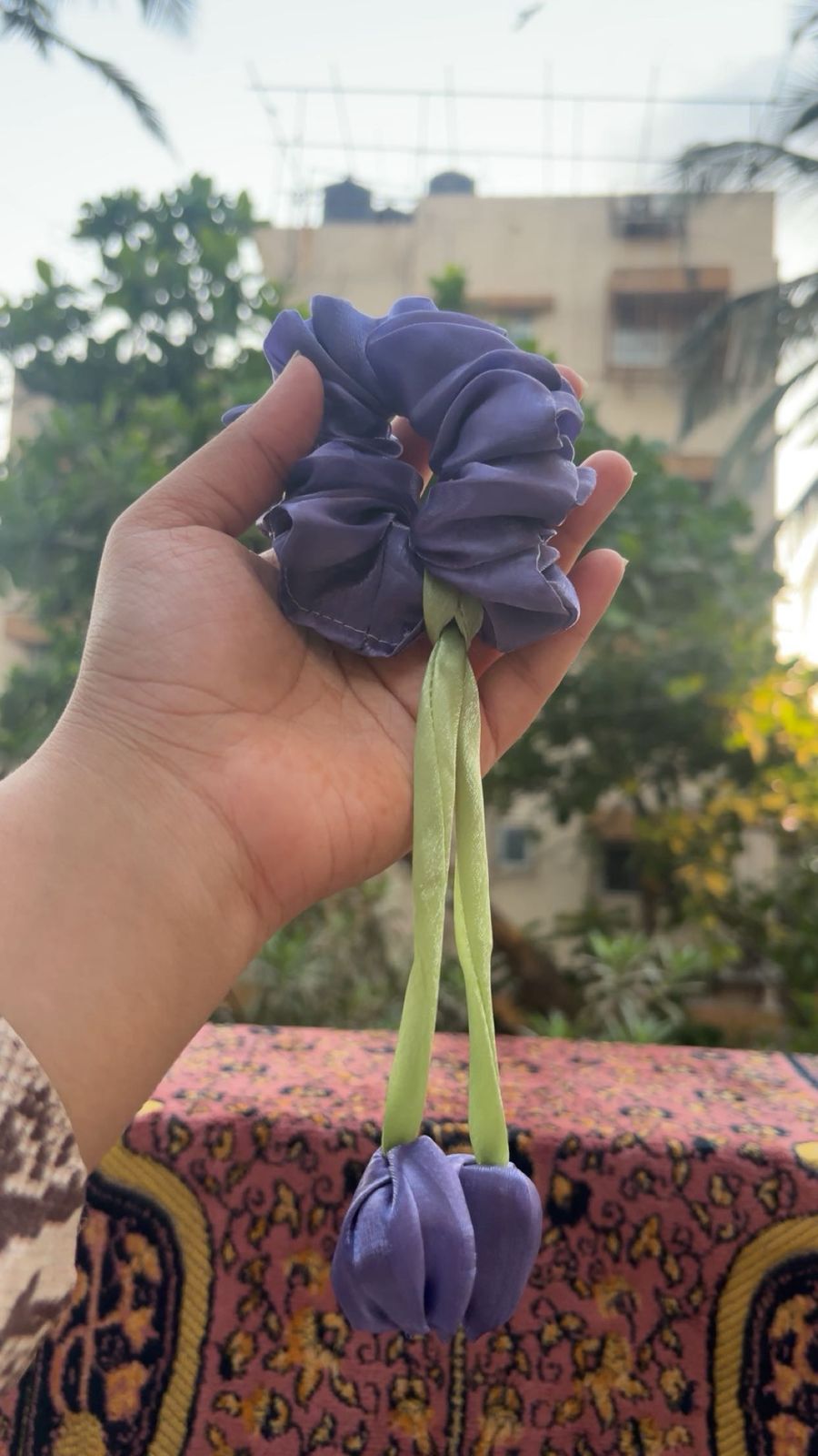 Purple fabric hair scrunchie with two light green ribbons being held in a hand outdoors. Used to tie hair.